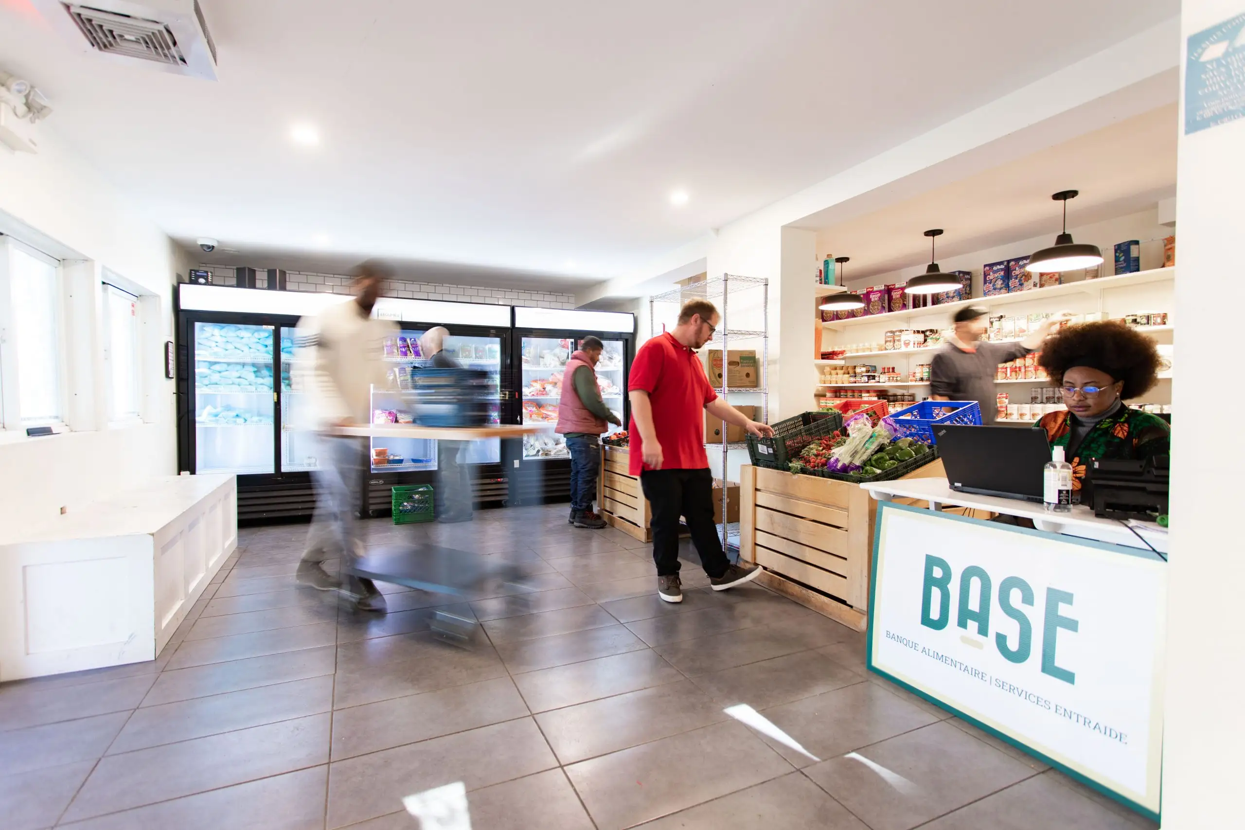 Entrée_66Eddy Photo dans laquelle on apperçoit Alex qui choisit des légumes dans le bacs à légumes, Carinne est au bureau derrière l'orgdianteur pour l'intervention sociale et en flou Nathan prépare un dépannage alimentaire au 66 rue Eddy à Gatineau